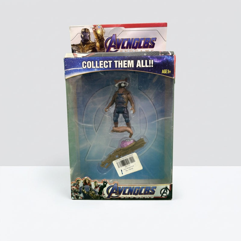 Blister card front with the raccoon-headed figure, its blaster accessory and the Age 3+ strip
